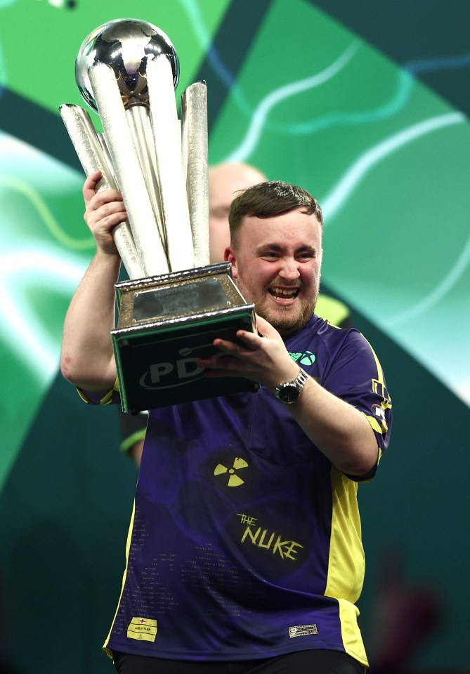 Luke Littler holding up a trophy after winning a darts championship.