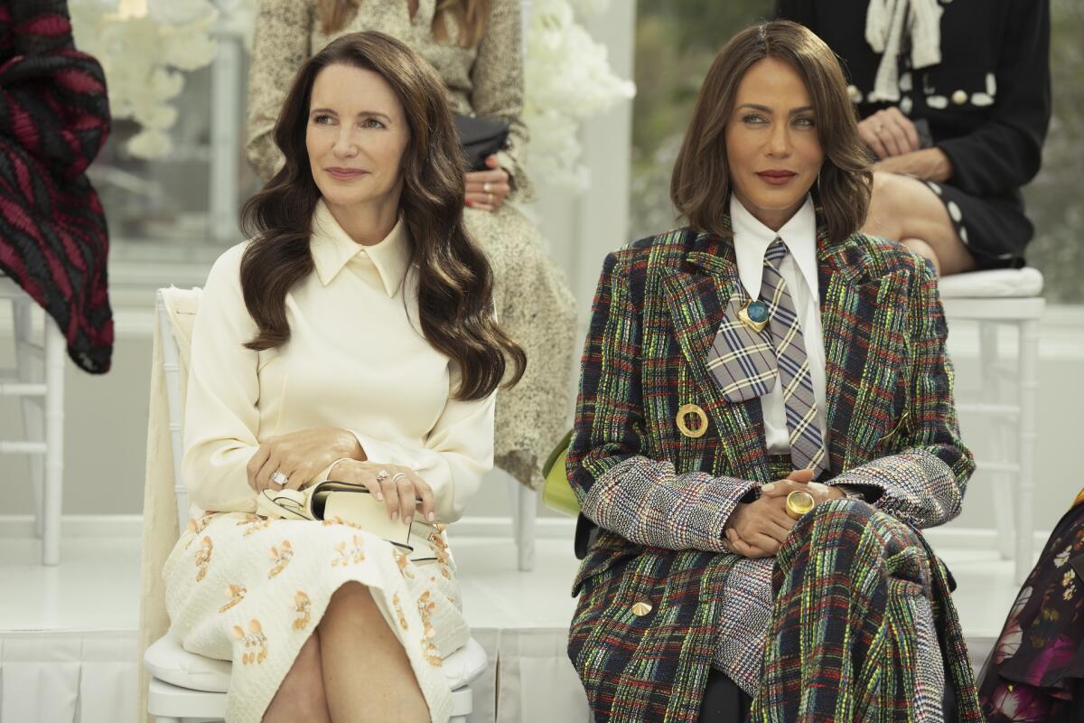 A woman in a white top and skirt sits next to a woman in a plaid coat.