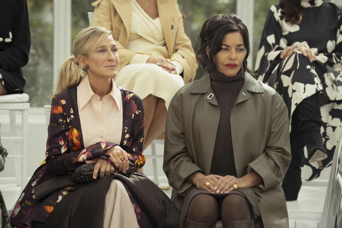 A woman in a dark floral coat and pink dress sits next a woman in a grey coat and brown turtleneck dress.