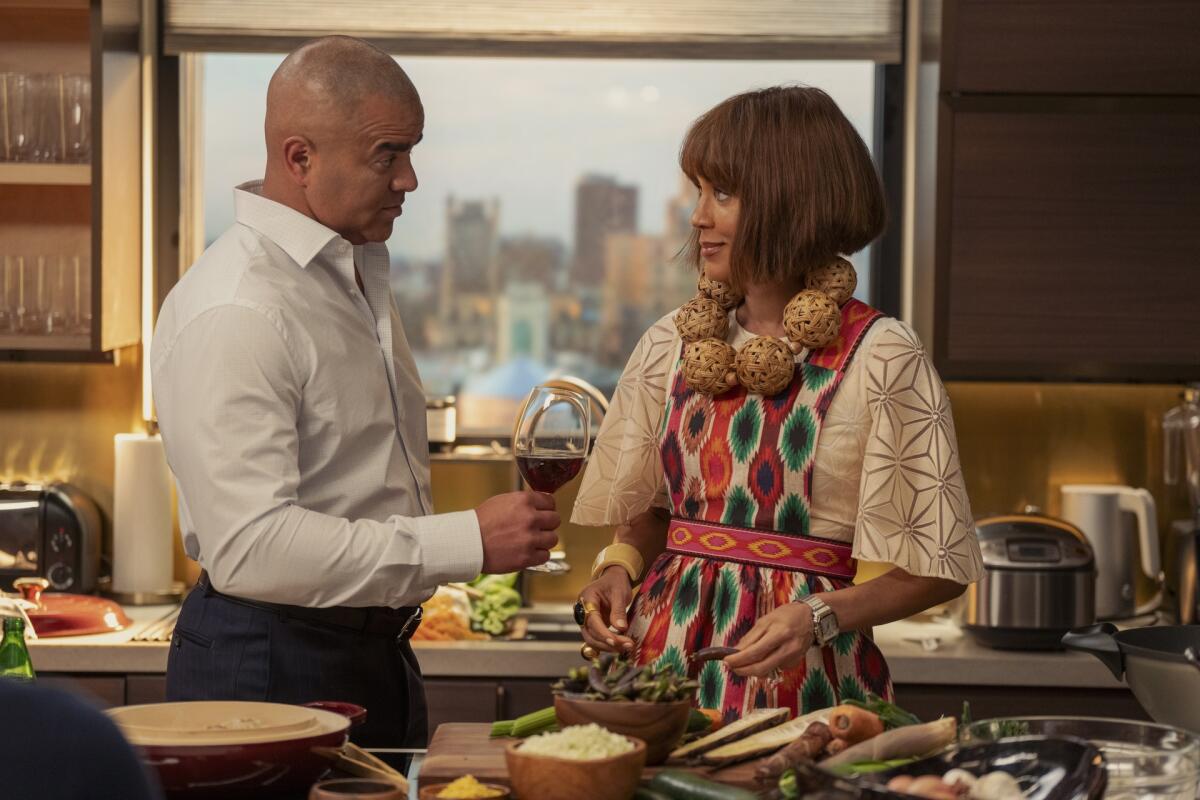 A man in a white shirt holds a wine glass toward a woman in a white dress, patterned apron and large necklace.