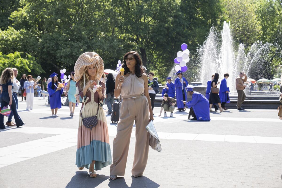 Two women eating ice cream as they walk through a park. A large fountain is in the background.