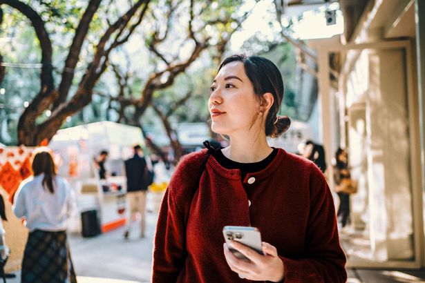 Young Asian woman using smartphone while enjoying a walk and exploring the city in street market. She is sightseeing and strolling along the street with Chinese New Year vibes.