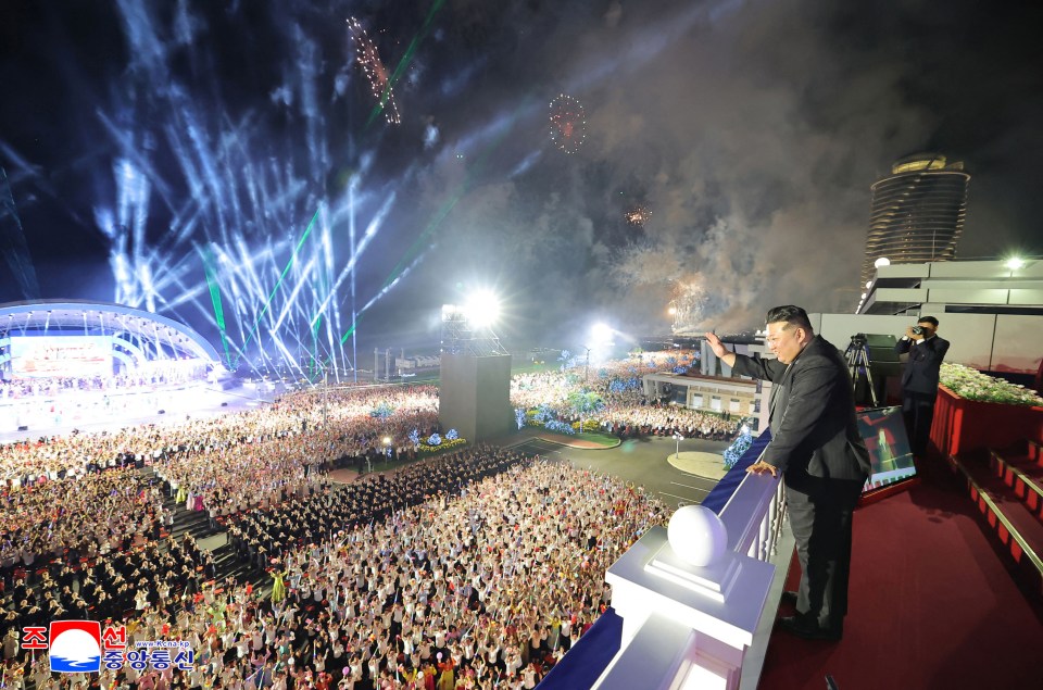 Kim Jong-un waving to a large crowd at a nighttime event with fireworks.