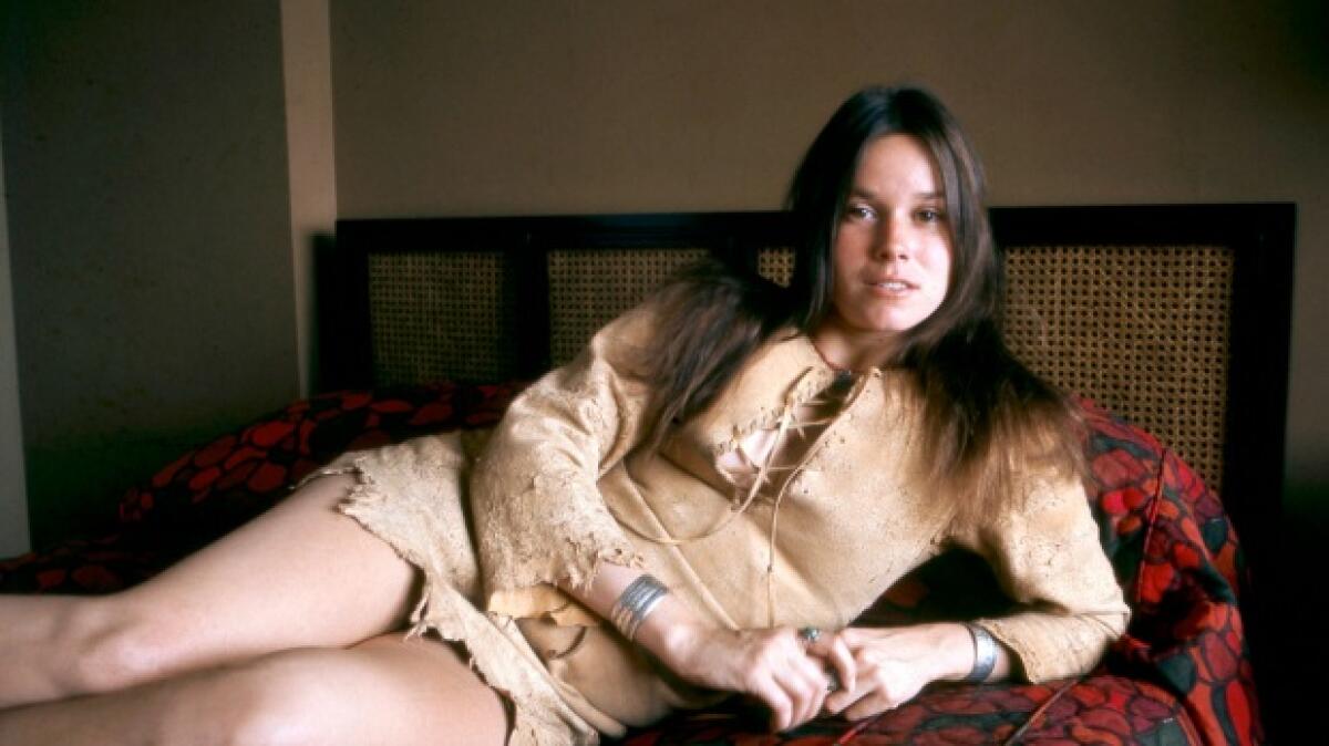 A woman in a beige dress reclines and looks at the lens.