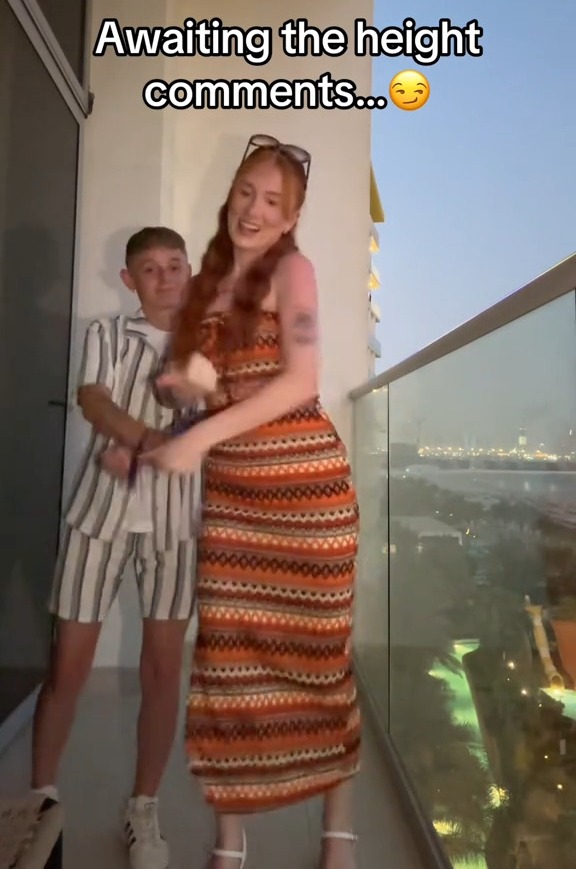 Video still of a couple on a balcony; the woman is significantly taller than the man.