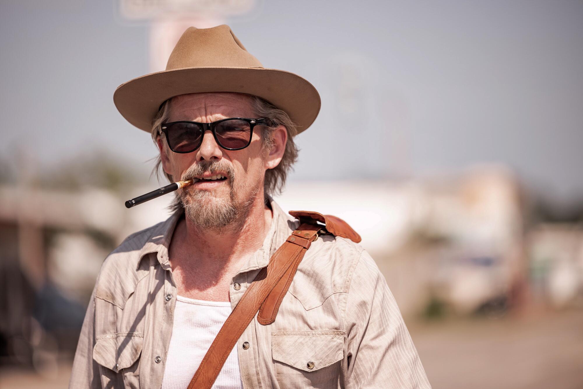 A man in a tan hat and sunglasses with a cigarillo between the side of his lips.