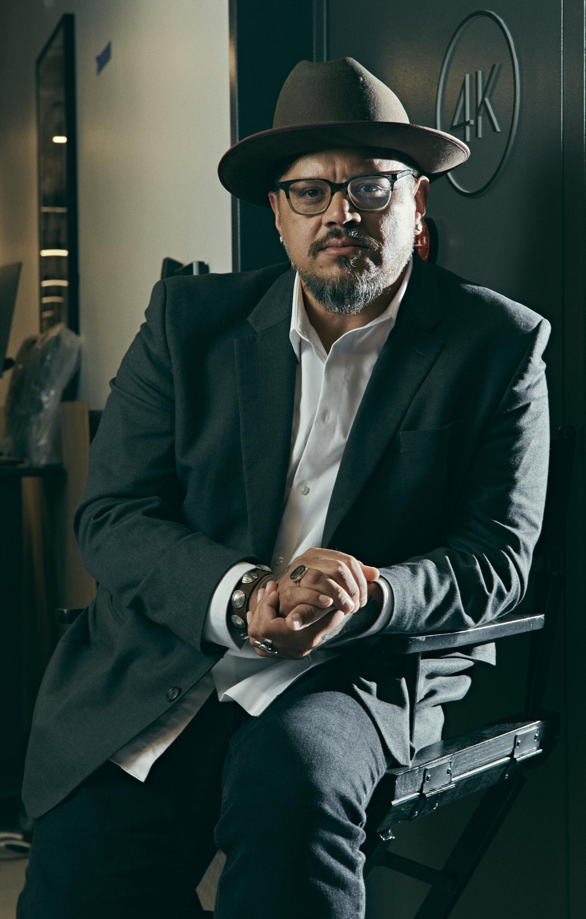 A man in a hat and glasses sits in a black directors chair.