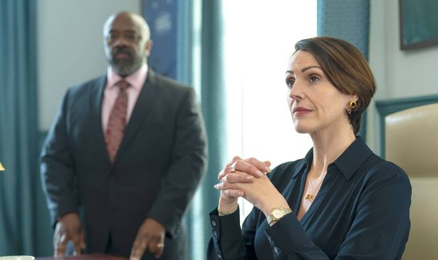 A man in a suit stands by a woman at a desk