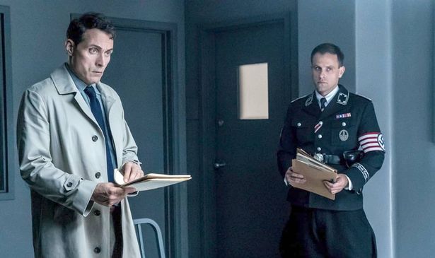 A man with a clipboard and a Nazi stand in a room