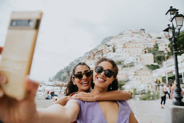 Two confident young woman stop in a street in Italy. They pose together as they take a selfie on a mobile smart phone. The famous Positano landscape is visible behind them.
