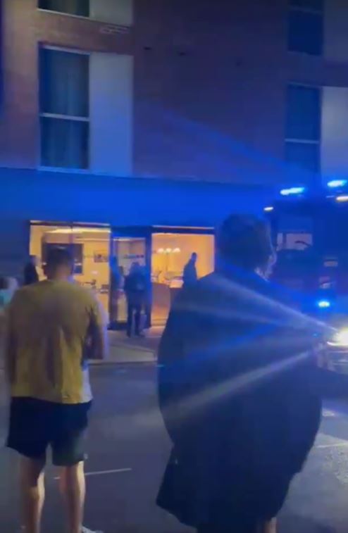 People standing outside a building at night as emergency lights flash.