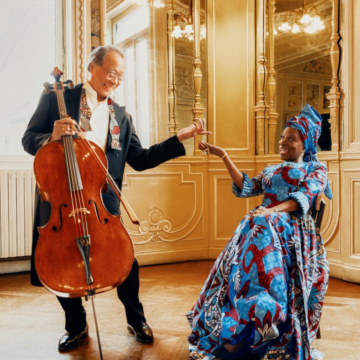 A man standing with a cello and a seated woman wearing a blue dress and matching headscarf gesture toward one another.