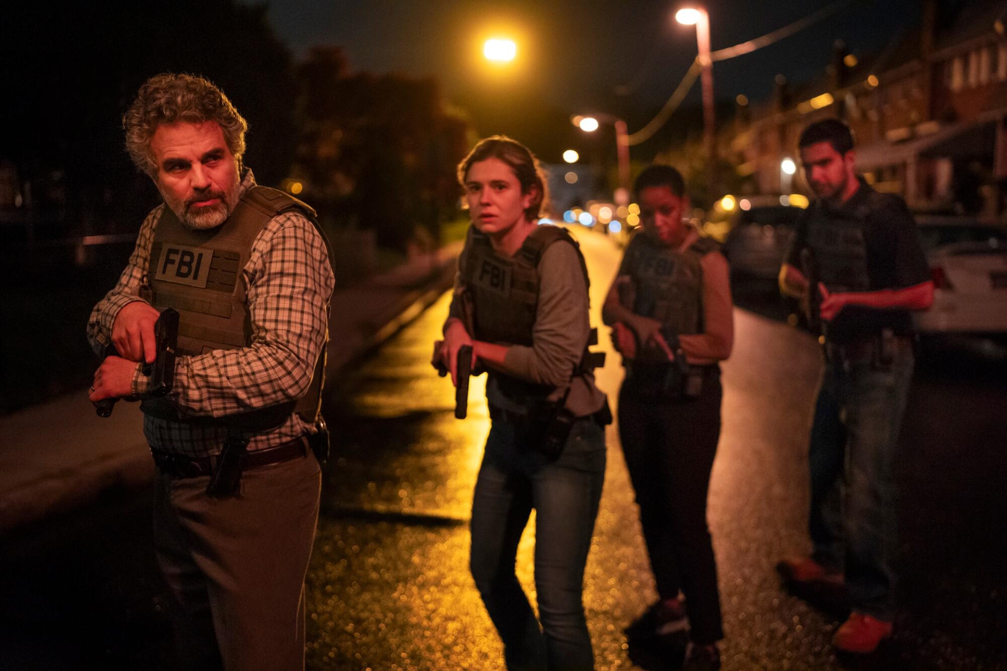 Four people in kevlar vests stand in a road with weapons drawn.