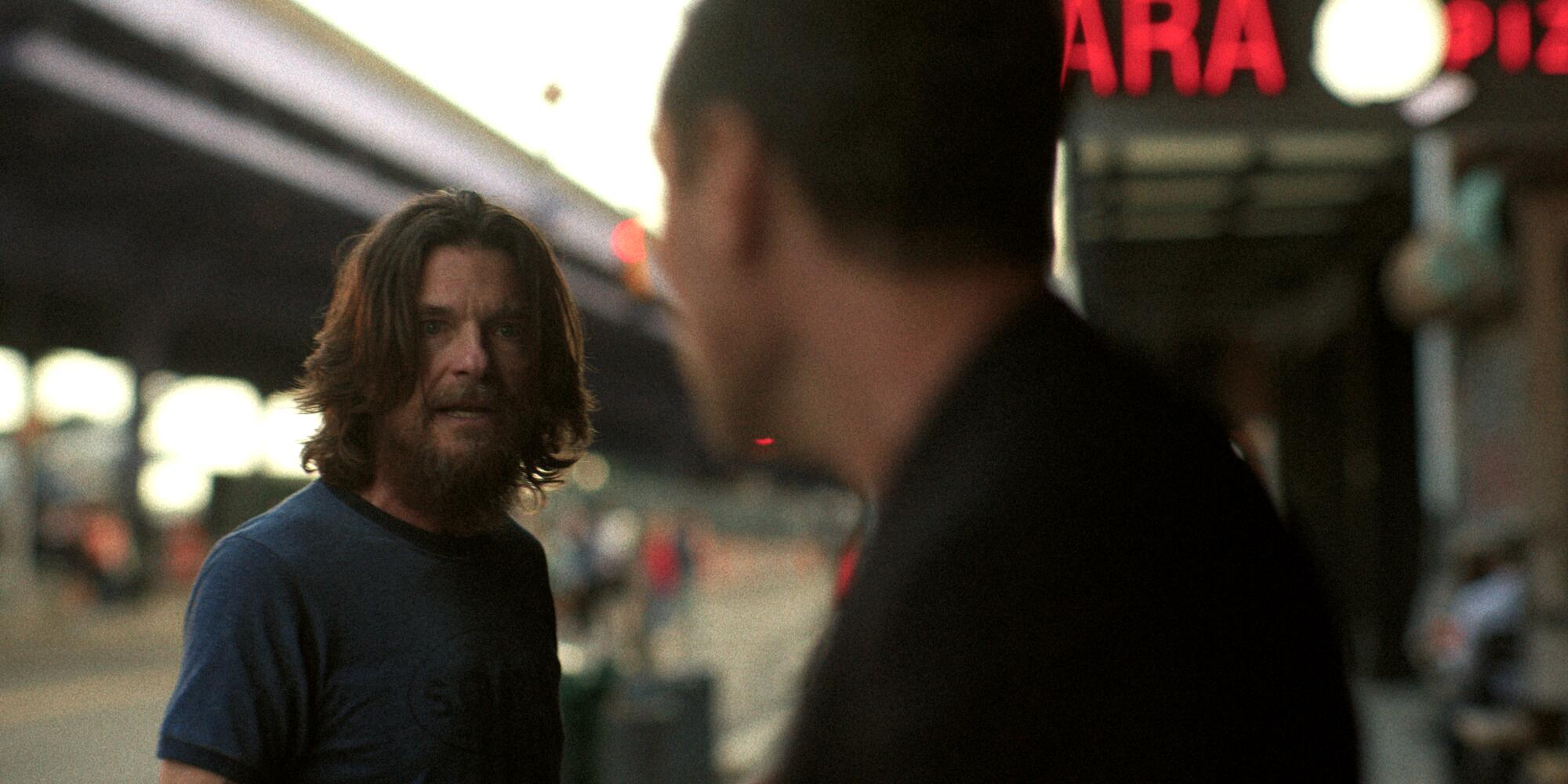 A man with a beard and shoulder-length shaggy hair looks at a man as they stand outside near a roadway.