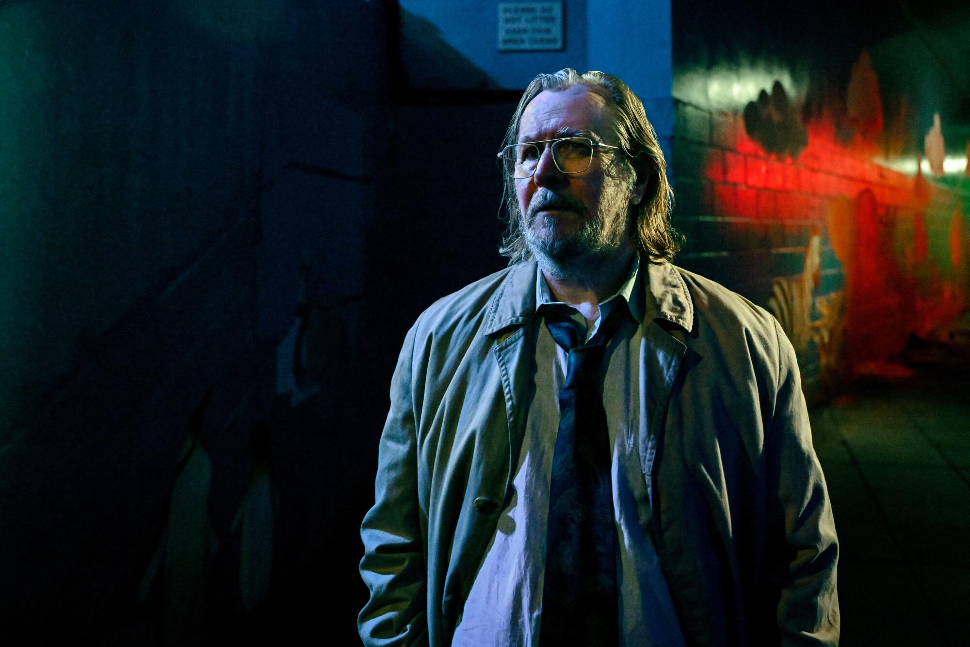 A man with glasses in a loosened tie and jacket stands near a brightly colored wall in the dark.