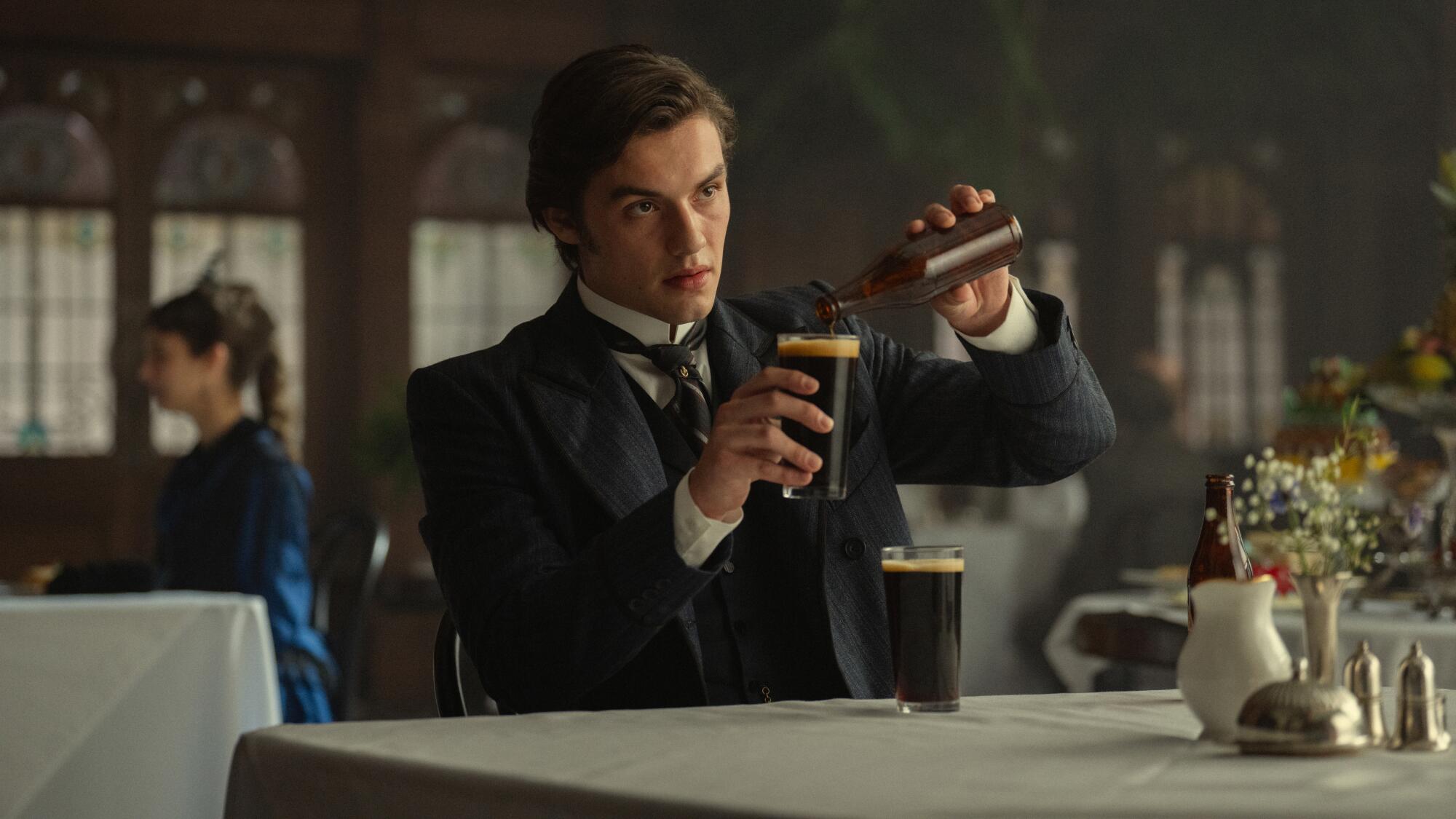 A man sitting at a table with a white linen cloth pours a dark beer into a glass from a beer bottle.