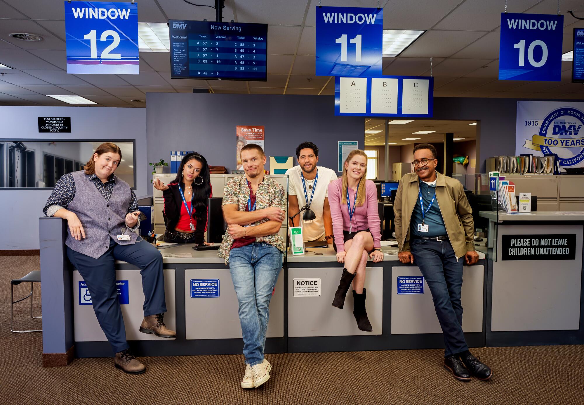 Six people sitting or standing at a grey counter with blue signs hanging above them.