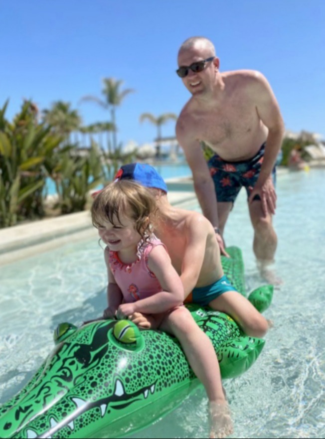 Two children riding an inflatable crocodile in a pool, with a man watching.