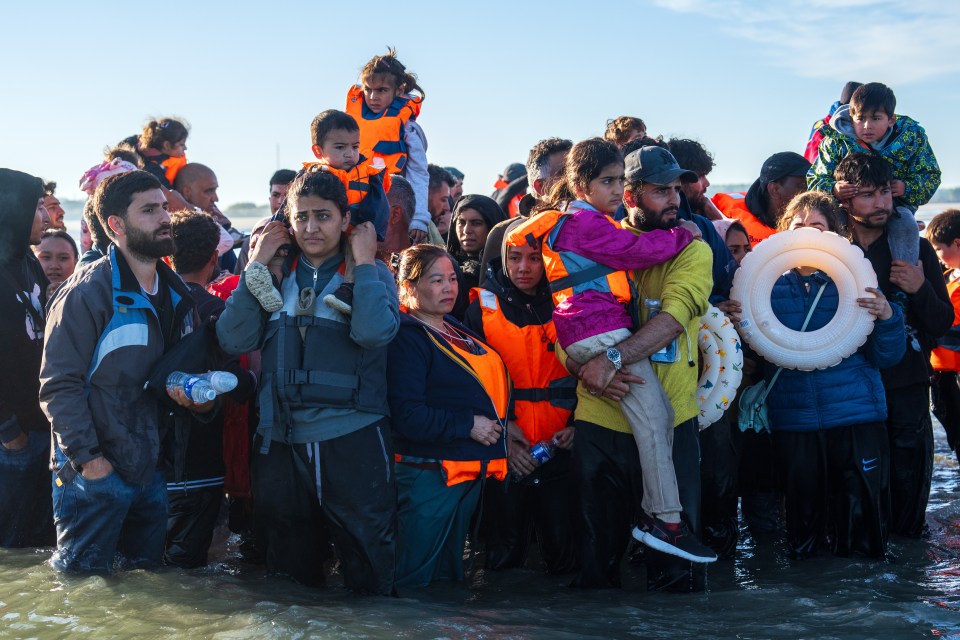 Migrant families in life vests wait in shallow water to board a boat.