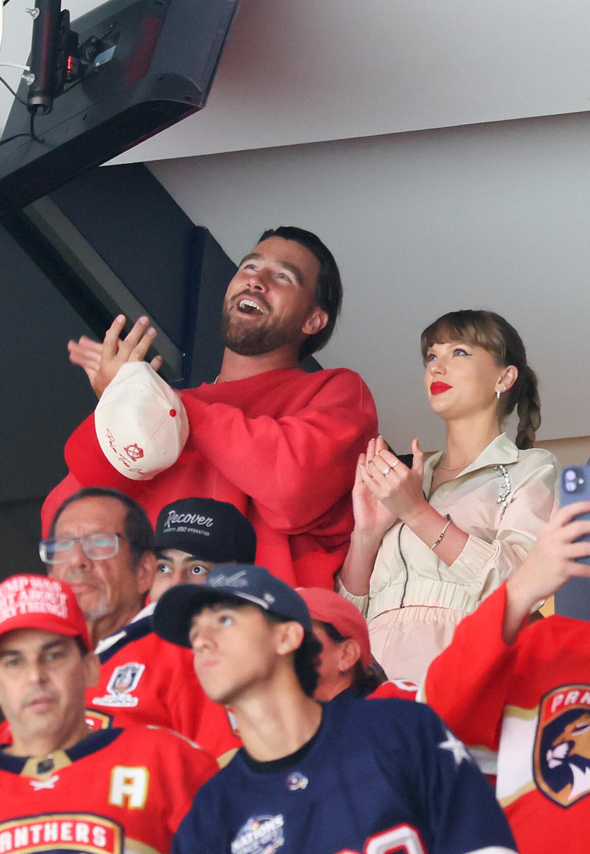 Travis Kelce laughs and claps and Taylor Swift claps in a luxury booth while watching an unseen hockey game