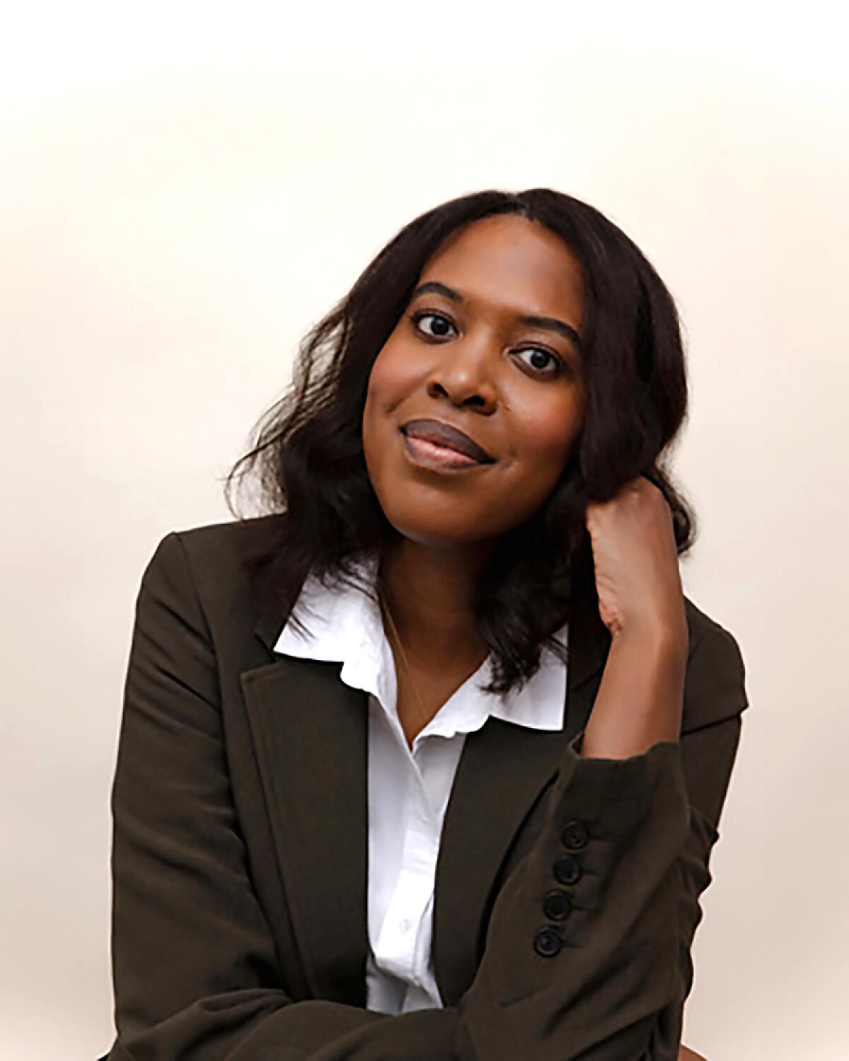 Aisha Muharrar, wearing a brown blazer and white collared shirt, rests her head on her fist.