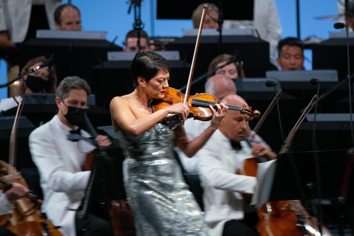 A female violinist in a sleeveless silver dress plays in front of an orchestra.  