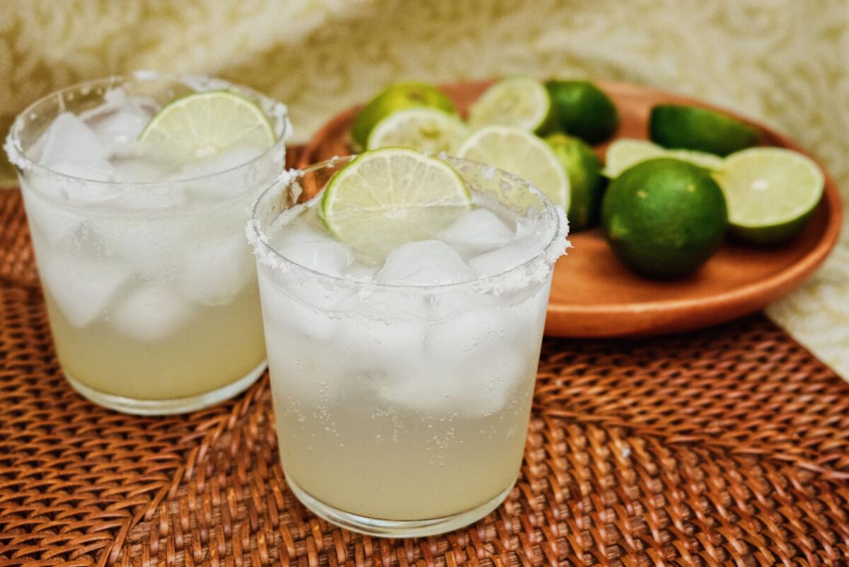Two suero drinks of lime and sparkling water on a brown textured placemat against green patterned fabric. Behind are limes.