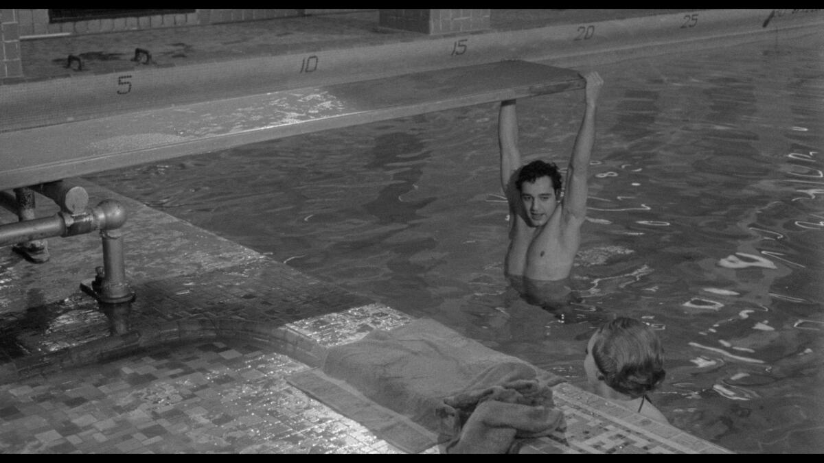 A young man hangs from a diving board in a pool.