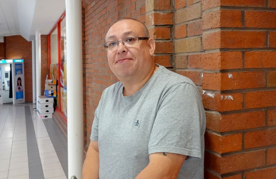 Man in grey shirt standing against brick wall.