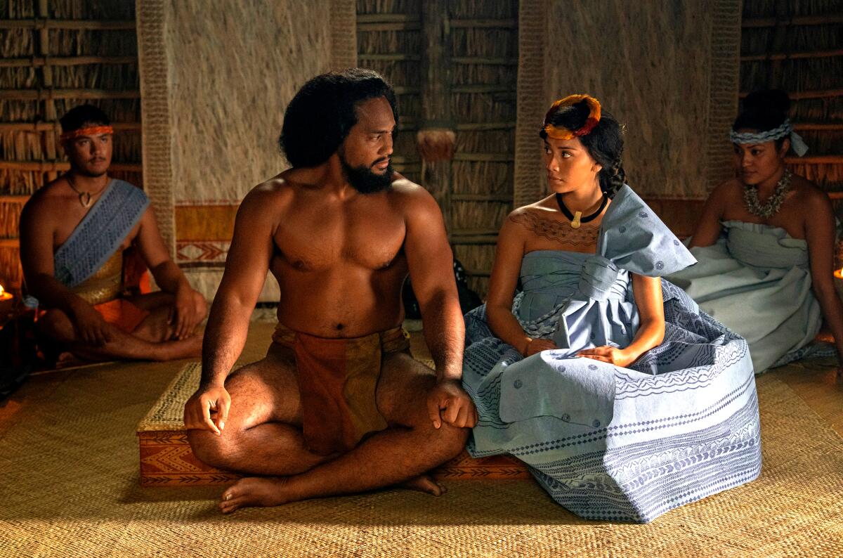 A man in a loin cloth sits cross legged next a woman in a voluminous blue dress.