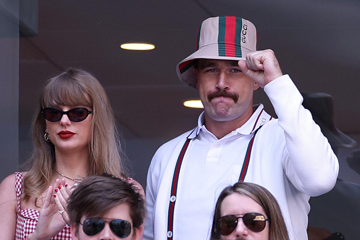 Taylor Swift wears shades and a mustached Travis Kelce wears a floppy hat and pumps his fist at a tennis match