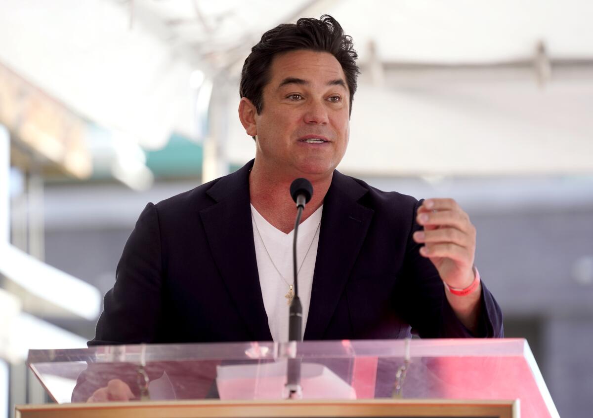 Dean Cain gestures with one hand while speaking into a small microphone at a lectern outdoors.