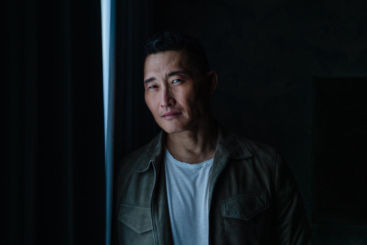 Actor Daniel Dae Kim, in a white shirt and olive green jacket, leans against a wall