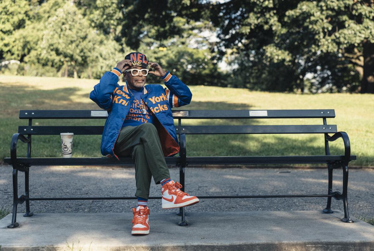 A man in Knicks gear and shades sits on a bench.