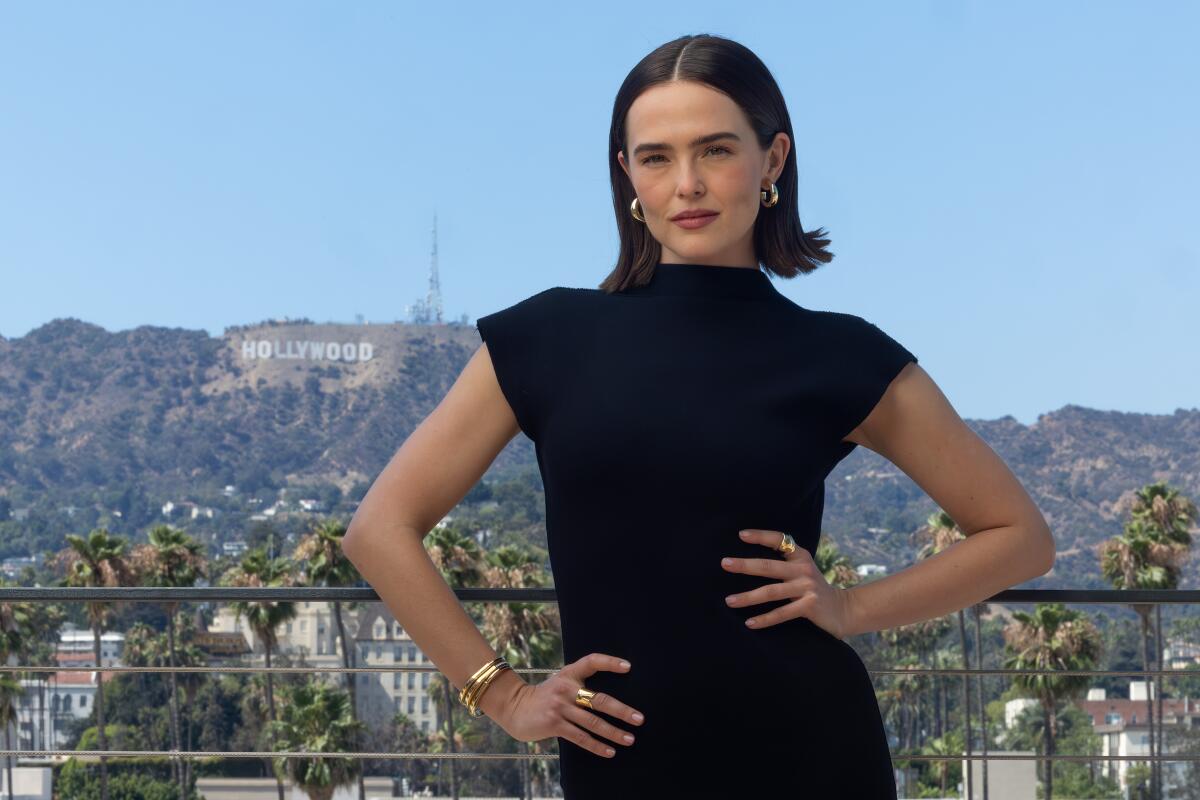 An actor in a dark dress poses in front of the Hollywood sign.