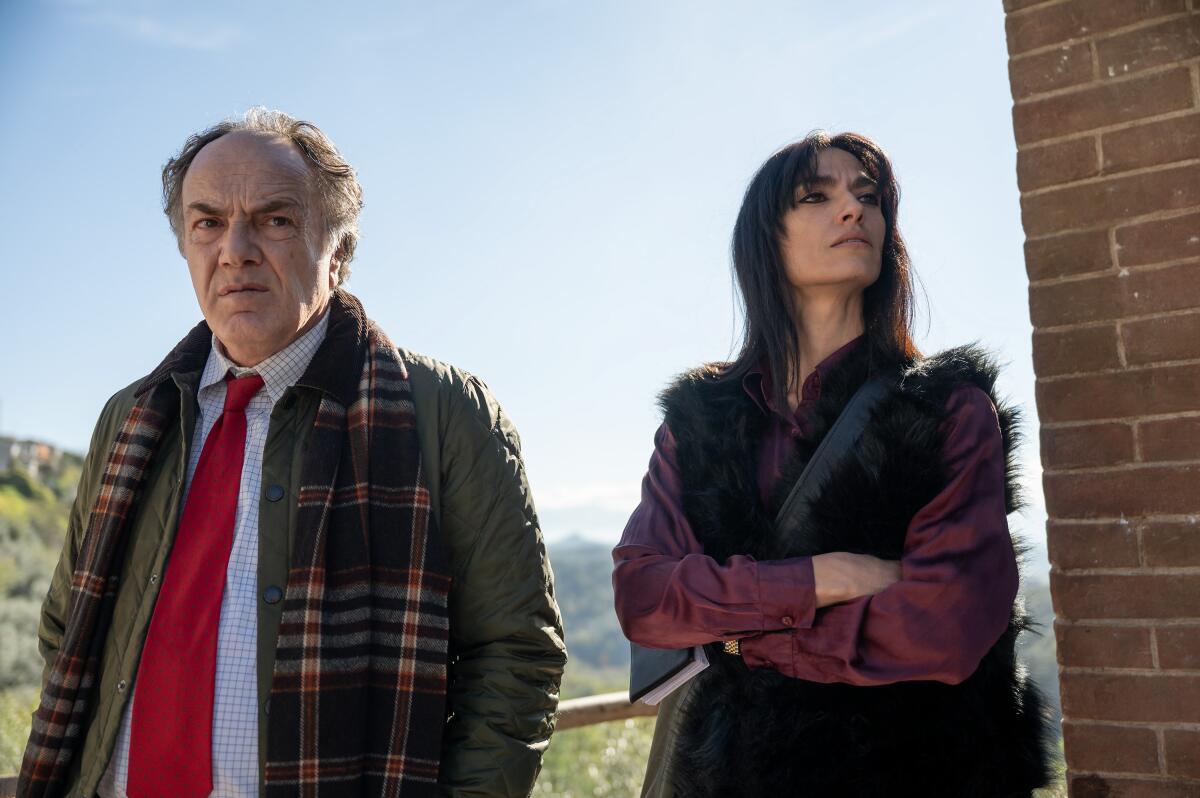 A man in a red tie and scarf around his shoulder stands next to a woman in a purple top and black vest who is looking away.