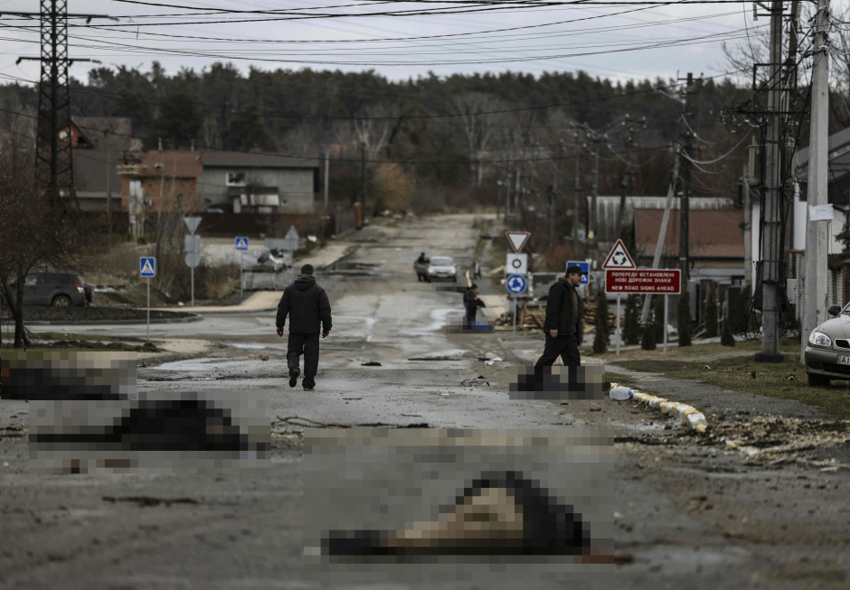 People walking past bodies lying on a damaged road.