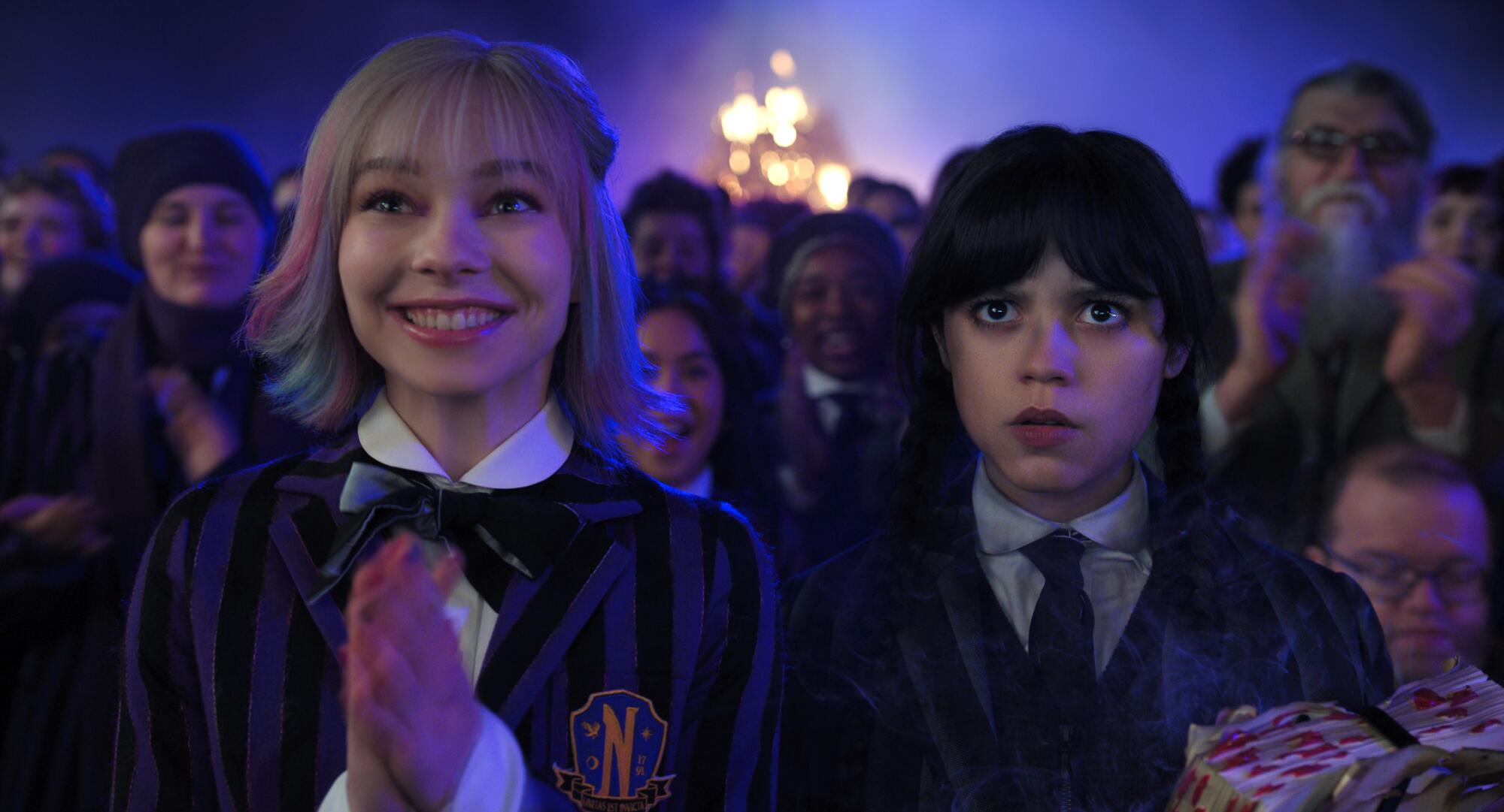 A smiling girl with colorful hair stands beside a girl with a flat expression