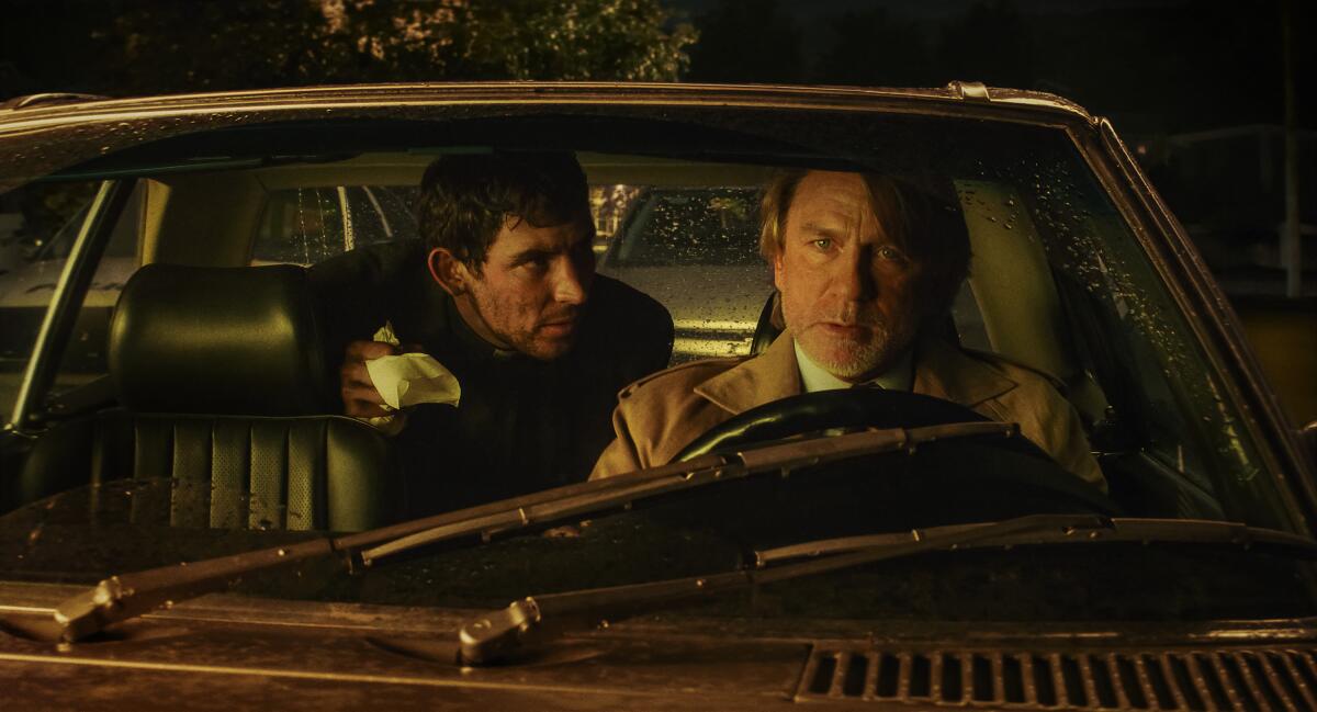 A man speaks to a worried driver from the backseat of a car.