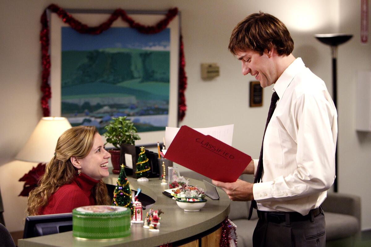 A woman sits at a desk while looking up at a man standing and holding a file folder