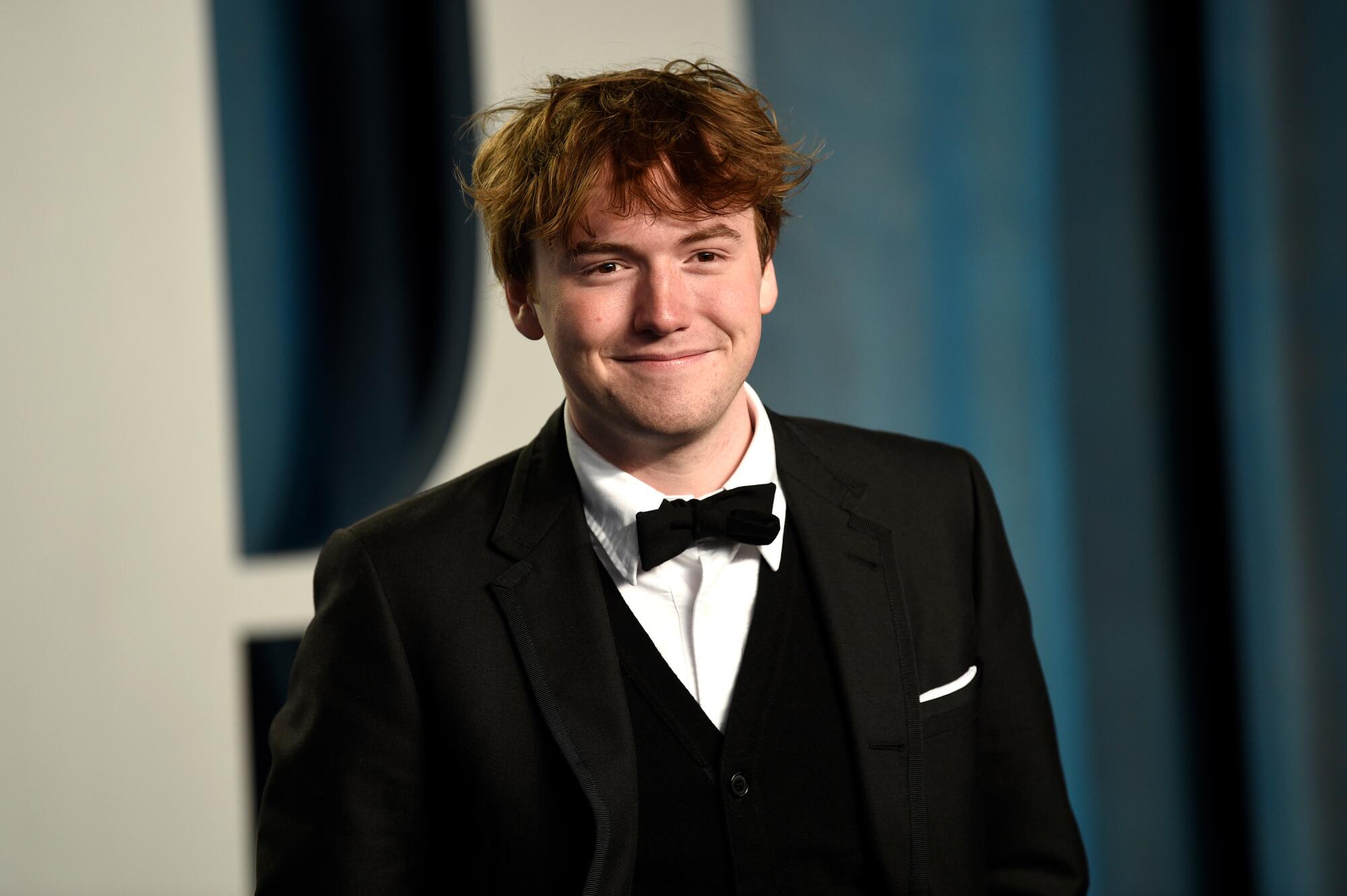 A young man in a tuxedo smiles on an arrival carpet.