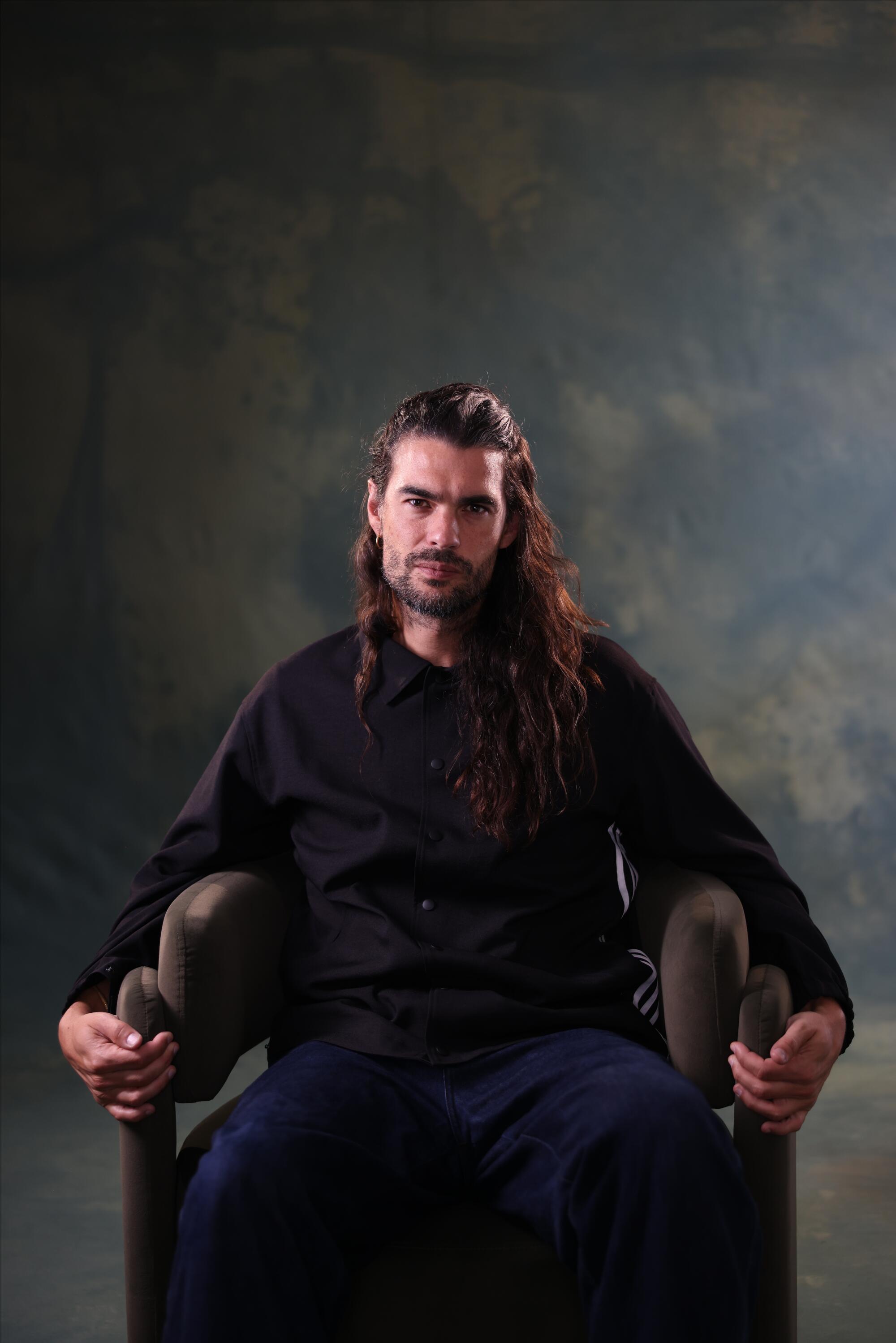 A bearded man with long hair sits in a chair.