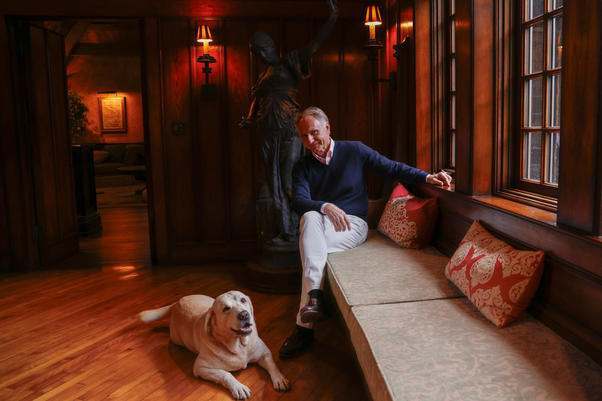 Dan Brown sits on a bench by a row of windows; his dog sits on the ground.