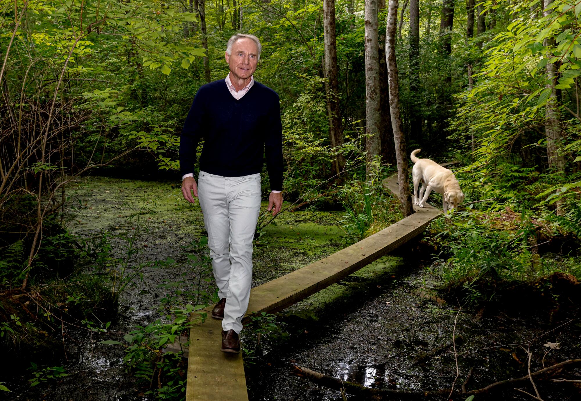 Dan Brown walks on a narrow walkway in the woods; his dog follows.