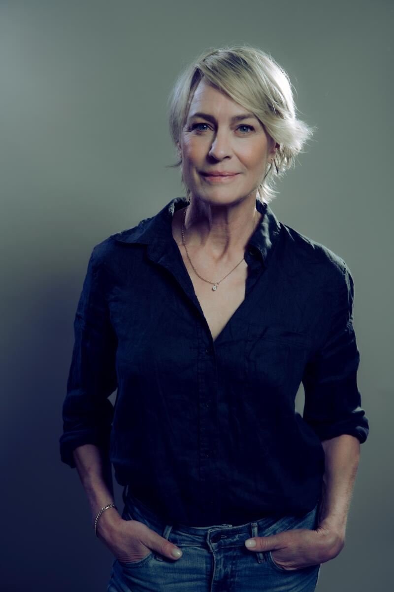 A blonde woman in a black shirt and jeans stands with her hands in her pockets.