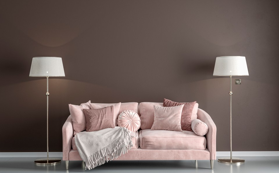 3D illustration of a pink velvet sofa with pillows and a throw blanket, flanked by floor lamps, against a brown wall.