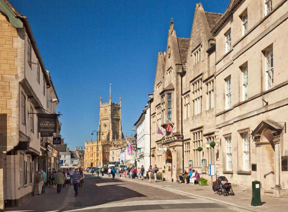 Street scene in Cirencester with shops and a church tower.