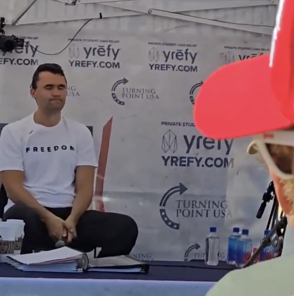 Charlie Kirk sitting at a table before a Turning Point USA backdrop.