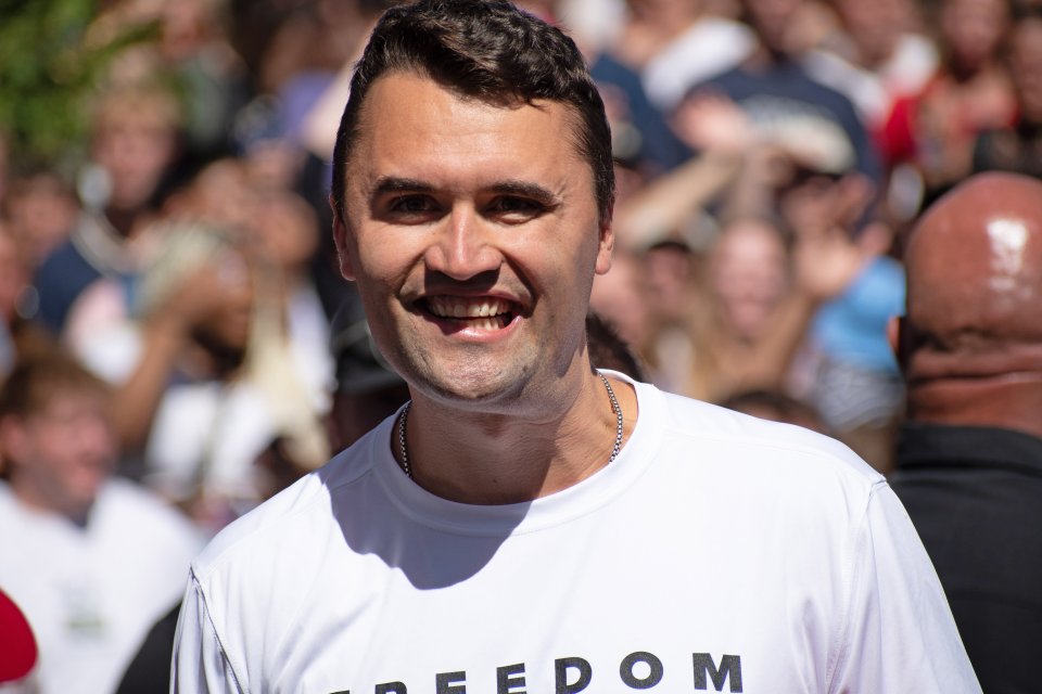 Charlie Kirk, wearing a "FREEDOM" t-shirt, smiles at a crowd.