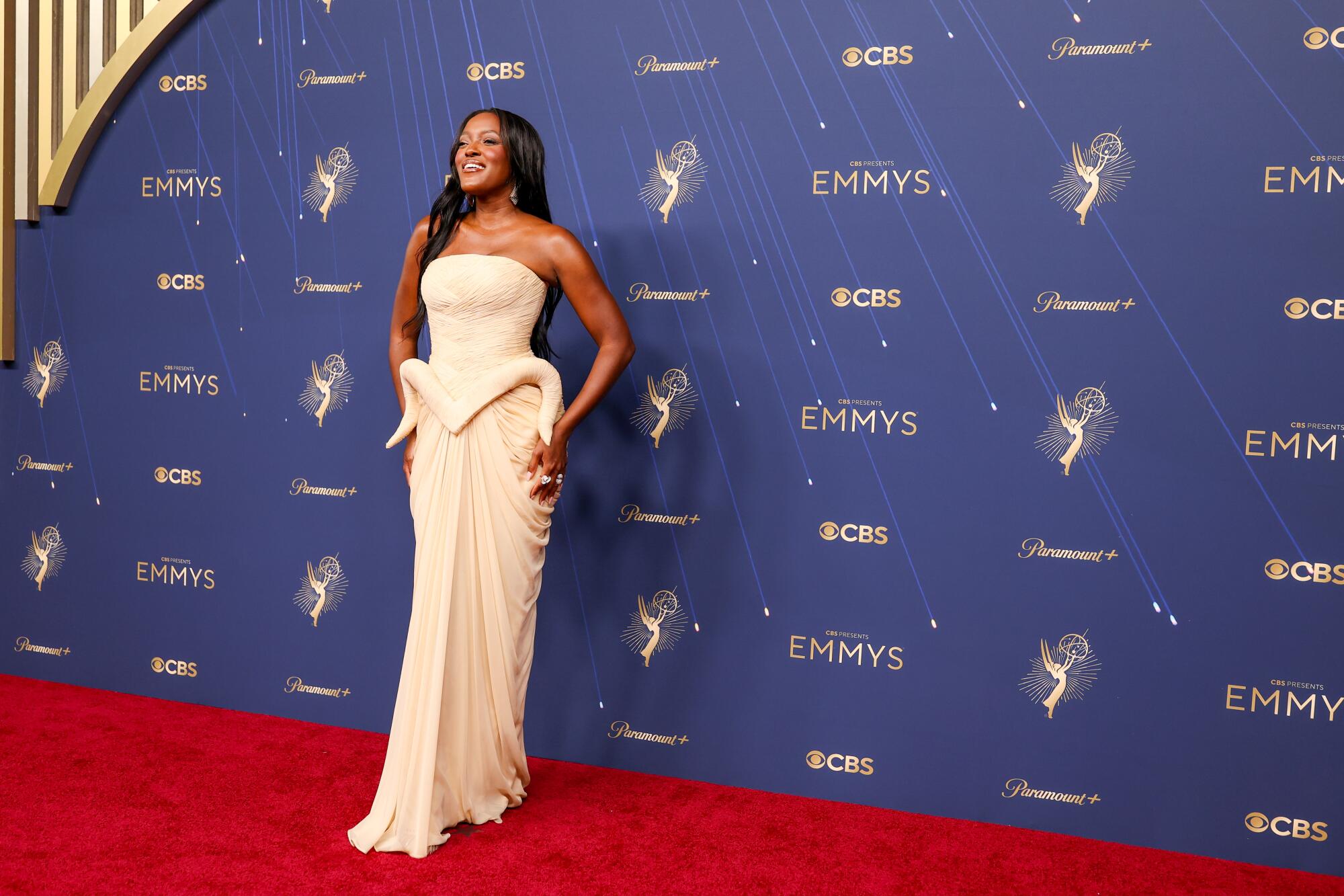 Krys Marshall wears a cream draped column dress with a horn-like embellishment at the waist.